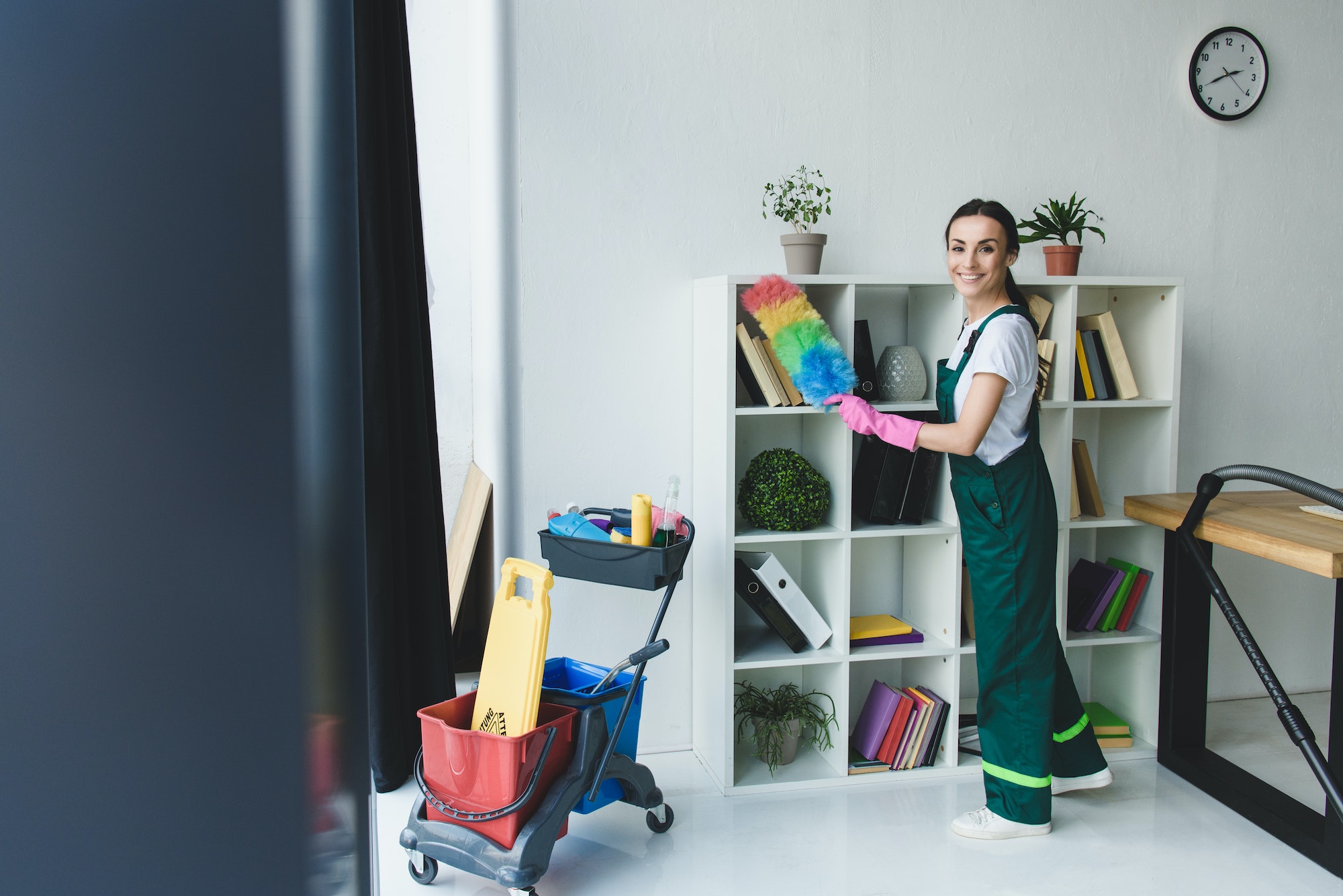 young cleaner holding duster and smiling at camera while cleaning shelves in office
